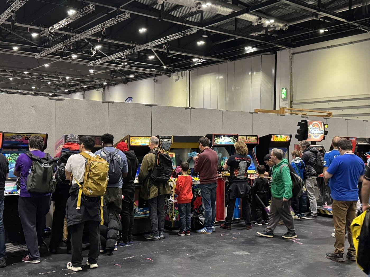 A row of classic arcade cabinets including Sega Daytona USA, Mortal Kombat, Rampage and Dance Dance Revolution SuperNOVA, with players of all ages at the controls