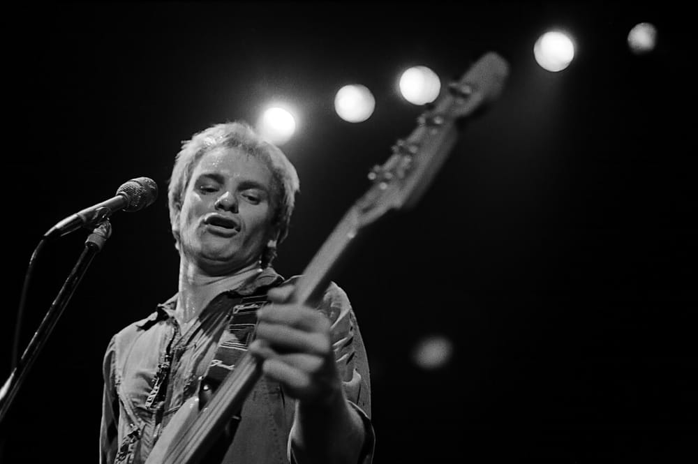 Black and white photo of a young Sting singing and playing bass on stage in 1979